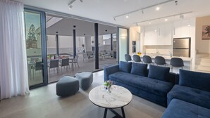 Una sala moderna con un sofá azul, una mesa de centro redonda y un comedor visible a través de puertas de vidrio.