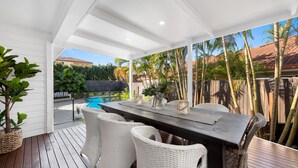 A covered patio with a pool, white wicker furniture, and a view of a tropical landscape.