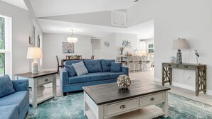 Uma sala de estar ampla com um sofá azul, uma mesa de centro de madeira e uma área de jantar ao fundo.
