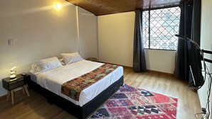 A bedroom with a wooden ceiling, a bed with a patterned bedspread, a nightstand with a lamp, a window with curtains, and a floor lamp. - BAMUKA HOSTEL (Bogotá)