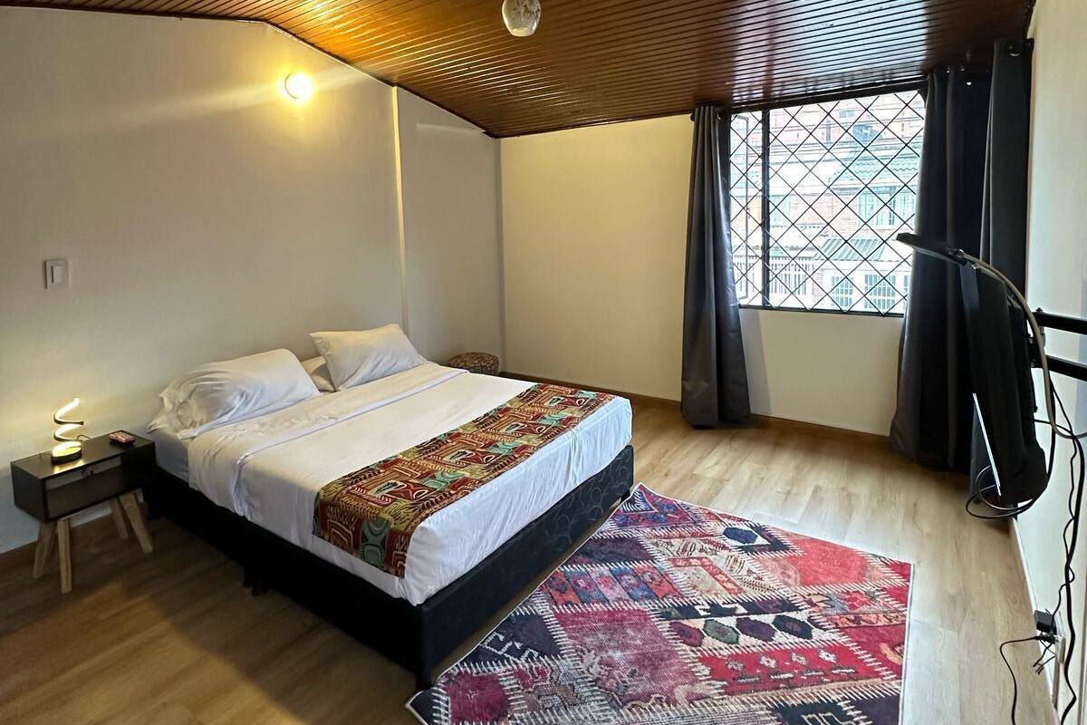 A bedroom with a wooden ceiling, a bed with a patterned bedspread, a nightstand with a lamp, a window with curtains, and a floor lamp.