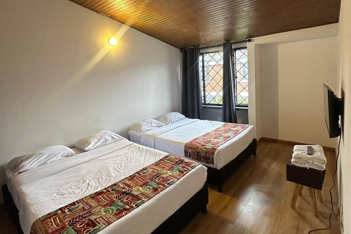 A room with two beds, wooden ceiling, a small table with a remote, and a window with curtains.