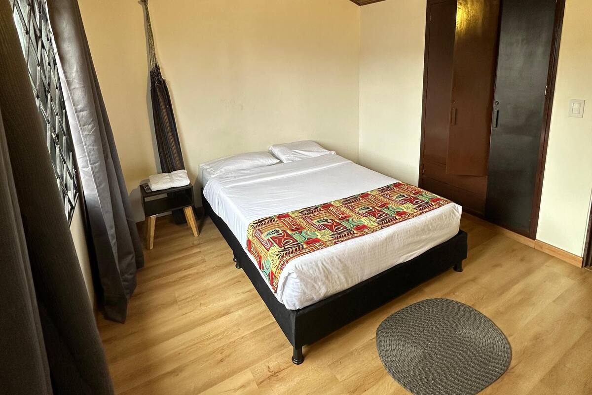 A bedroom with a wooden ceiling, a bed with a colorful blanket, a small bedside table, and a grey rug.