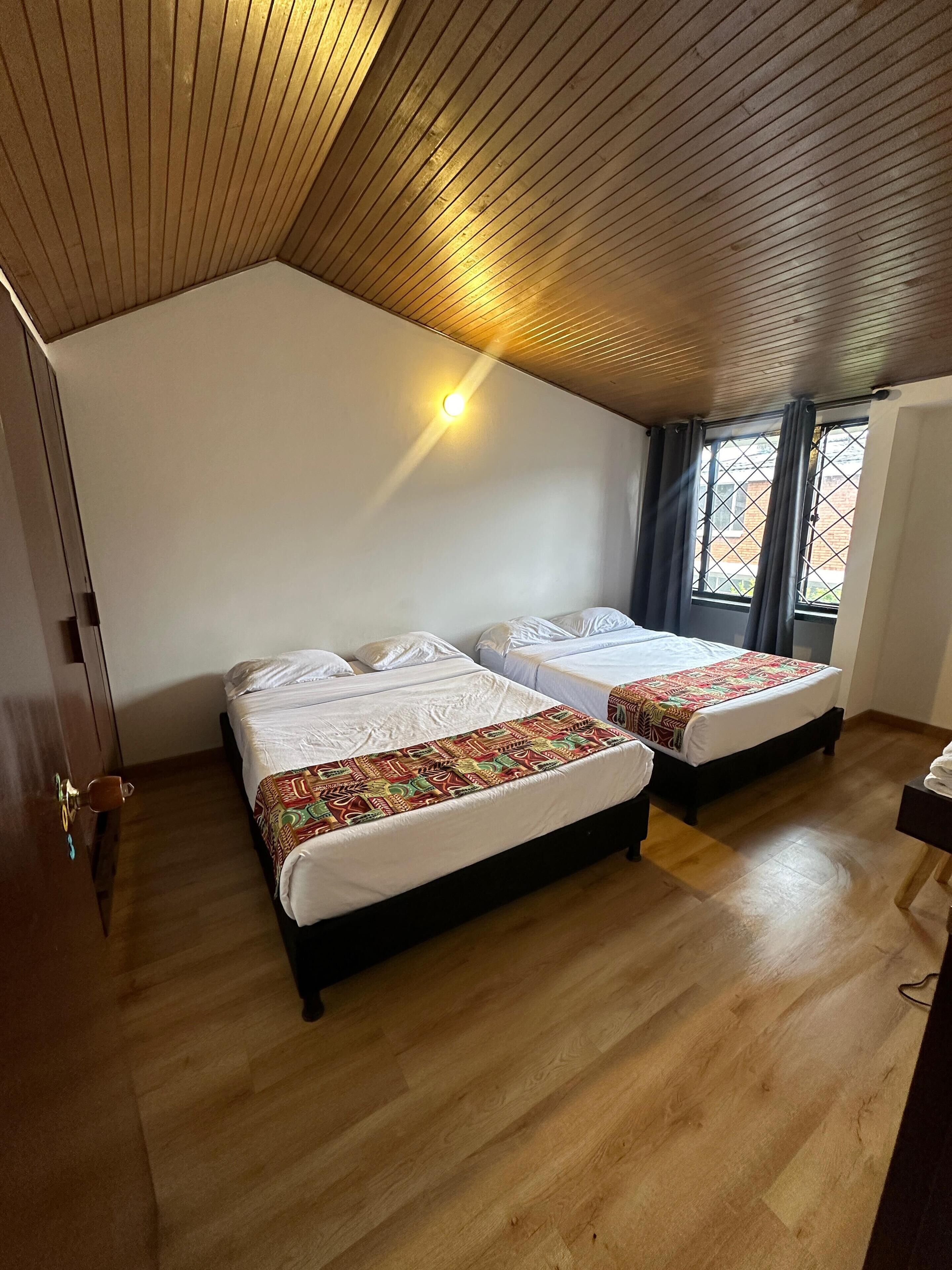 Habitación con dos camas, suelo de madera, techo inclinado, una ventana con cortinas y una lámpara fijada a la pared.