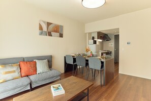A modern living room with a sofa, dining table, and chairs, a kitchen in the background, and a book on the coffee table. - AQUA RESIDENCE Sangenjaya by Tranova (Tokyo)