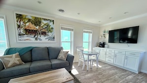 A living room with a gray sofa, a coffee table, a dining table with chairs, a kitchen area with white cabinets, a TV mounted on the wall, and a large painting of a beach scene. - New Waterfront❗️Pool, Epic Views/Corner Lot❤️Beaches near~BBQ Grill~Washer/Dryer (Jensen Beach)