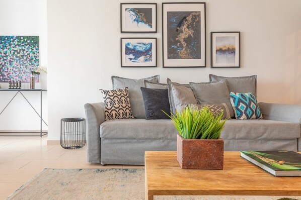 Ein modernes Wohnzimmer mit grauem Sofa, mehreren Kissen, einem kleinen Beistelltisch mit Pflanze und gerahmten Kunstwerken an der Wand.
