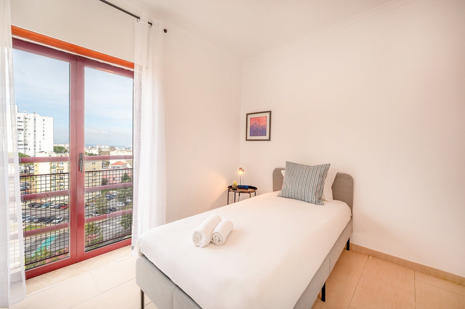 Een hotelkamer met een bed, een nachtkastje, een lamp, een raam met uitzicht op de stad en een schilderij aan de muur.