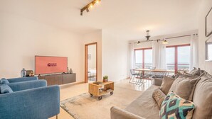 A modern living room with a sofa, a TV, a dining area with a table and chairs, and a view of the sea through large windows. - GuestReady - A relaxing retreat in Lisbon (Lisbon)