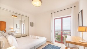 A modern hotel room with a bed, a desk, a chair, a lamp, a mirror, and a view of the sea through a glass door. - GuestReady - A relaxing retreat in Lisbon (Lisbon)