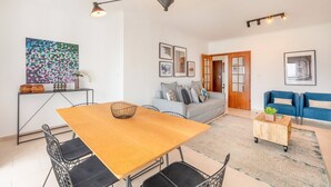 A modern living room with a wooden dining table, black wire chairs, a grey sofa, and a blue sofa. There are framed pictures on the walls and a colorful abstract painting above the sofa. - GuestReady - A relaxing retreat in Lisbon (Lisbon)