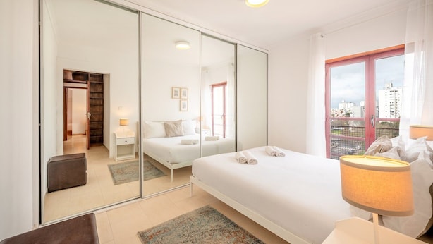 A modern hotel room with a large bed, a sitting area with a chair, a desk, and a large window offering a city view. - GuestReady - A relaxing retreat in Lisbon (Lisbon)