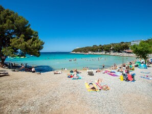 A beach with people sunbathing and swimming, clear blue water, and a tree-lined shoreline.