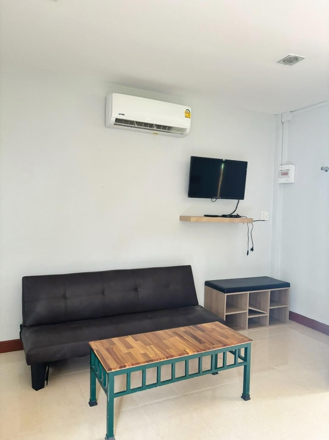 Una sala de estar con un sofá negro, una mesa de centro de madera, un televisor montado en la pared y un pequeño mueble de madera.