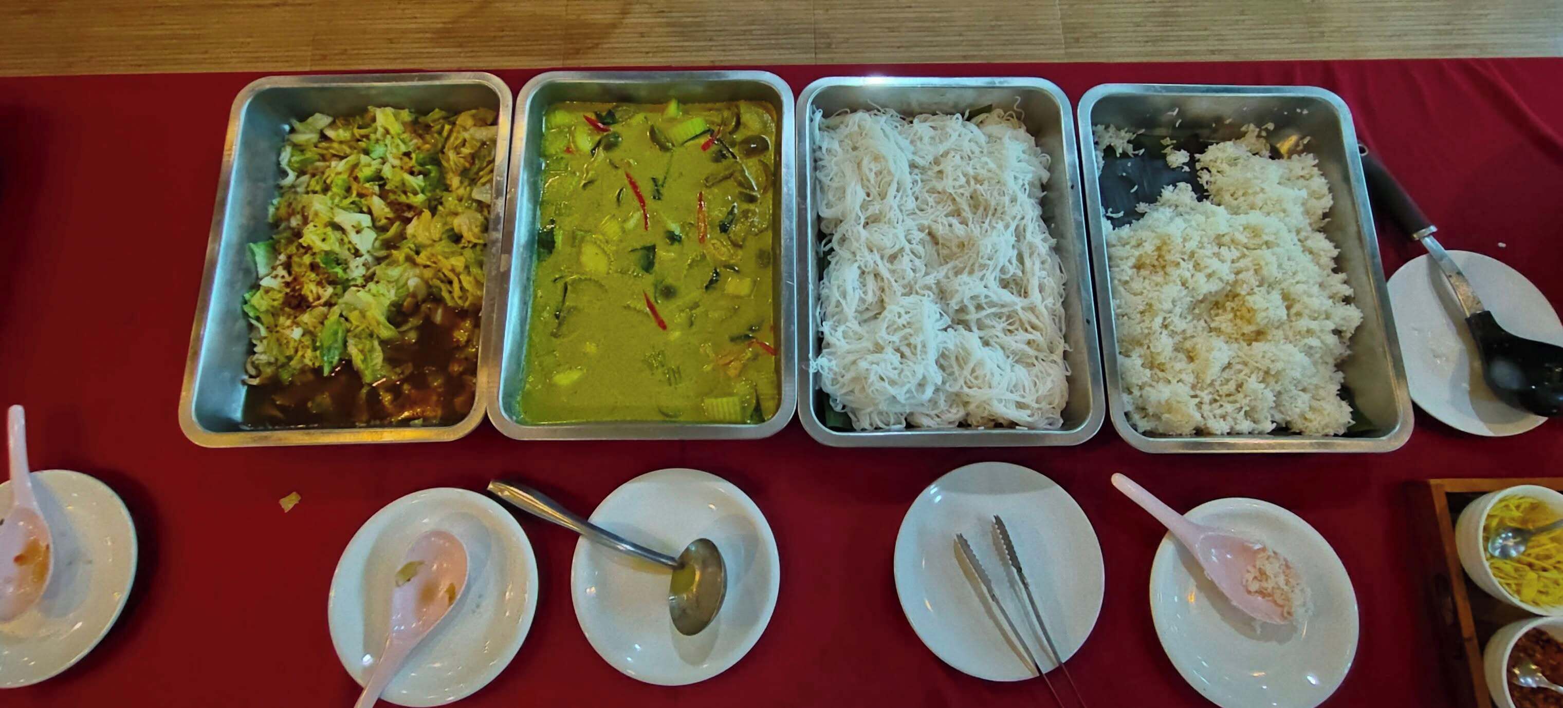 A buffet table with four metal trays containing different dishes, and several small plates with utensils.
