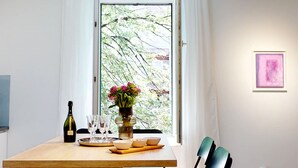 A dining table with a bottle, glasses, and a vase of flowers by a window with a view of trees.