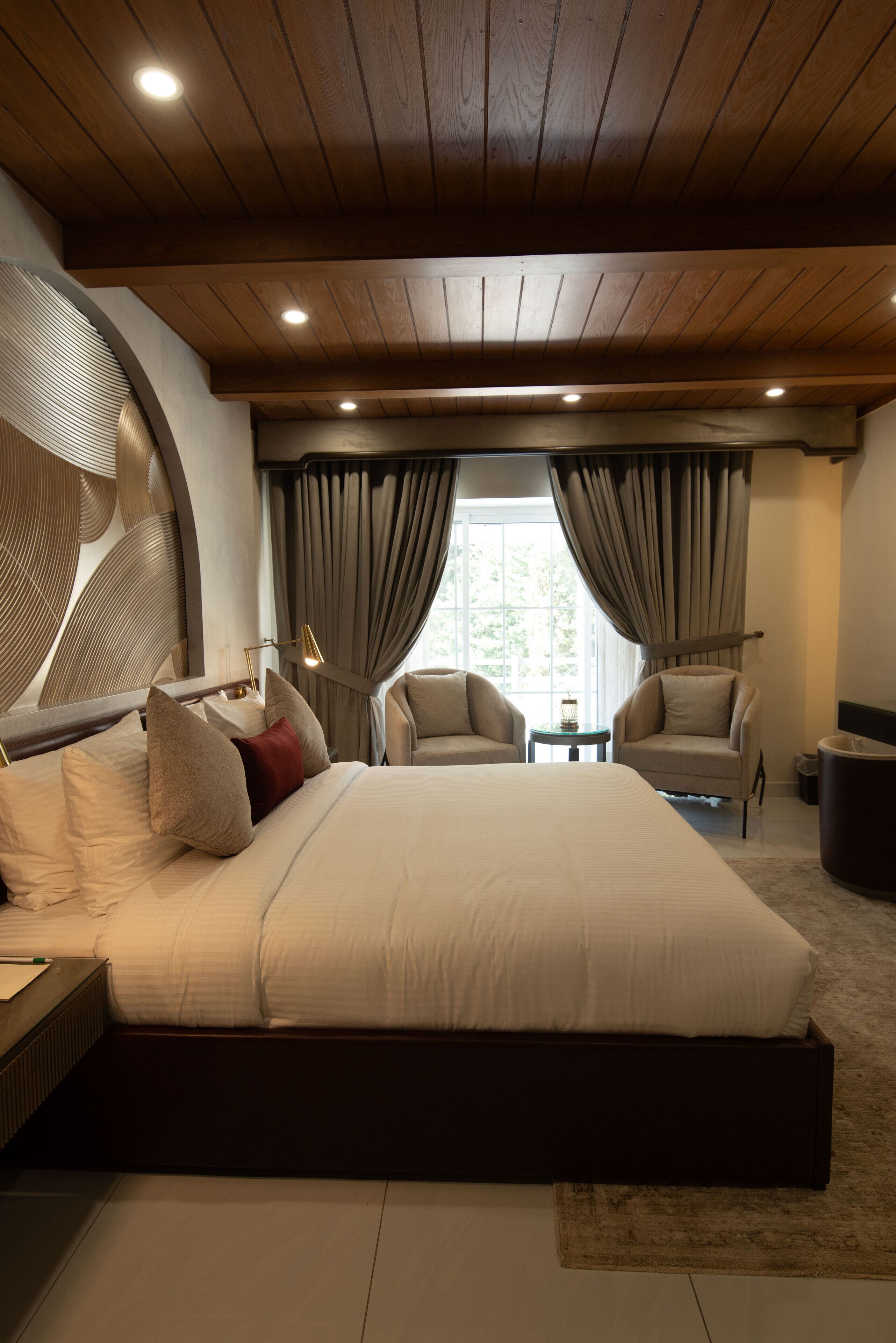 A bedroom with a large bed, two armchairs, a small table, and a view of the outdoors.