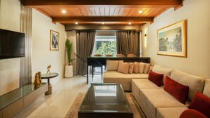 Un salon moderne avec un plafond en bois, un grand téléviseur à écran plat, un canapé beige agrémenté de coussins rouges, une table basse en verre et un coin repas avec une table et des chaises noires.
