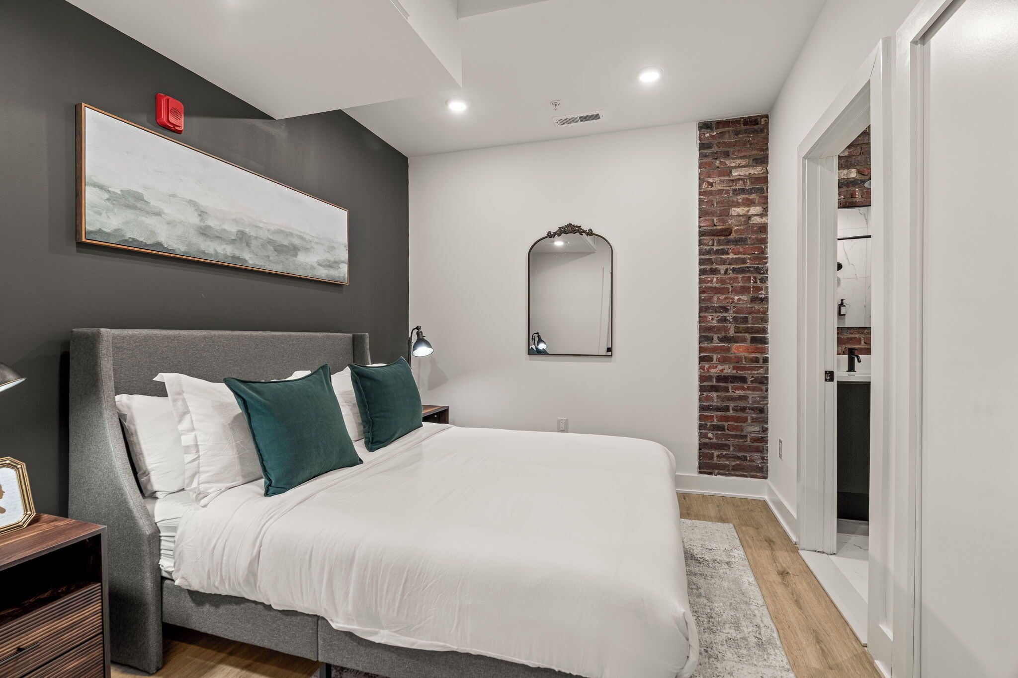 Une chambre moderne avec un grand lit, une table de chevet, un miroir et un mur d’accent en briques.