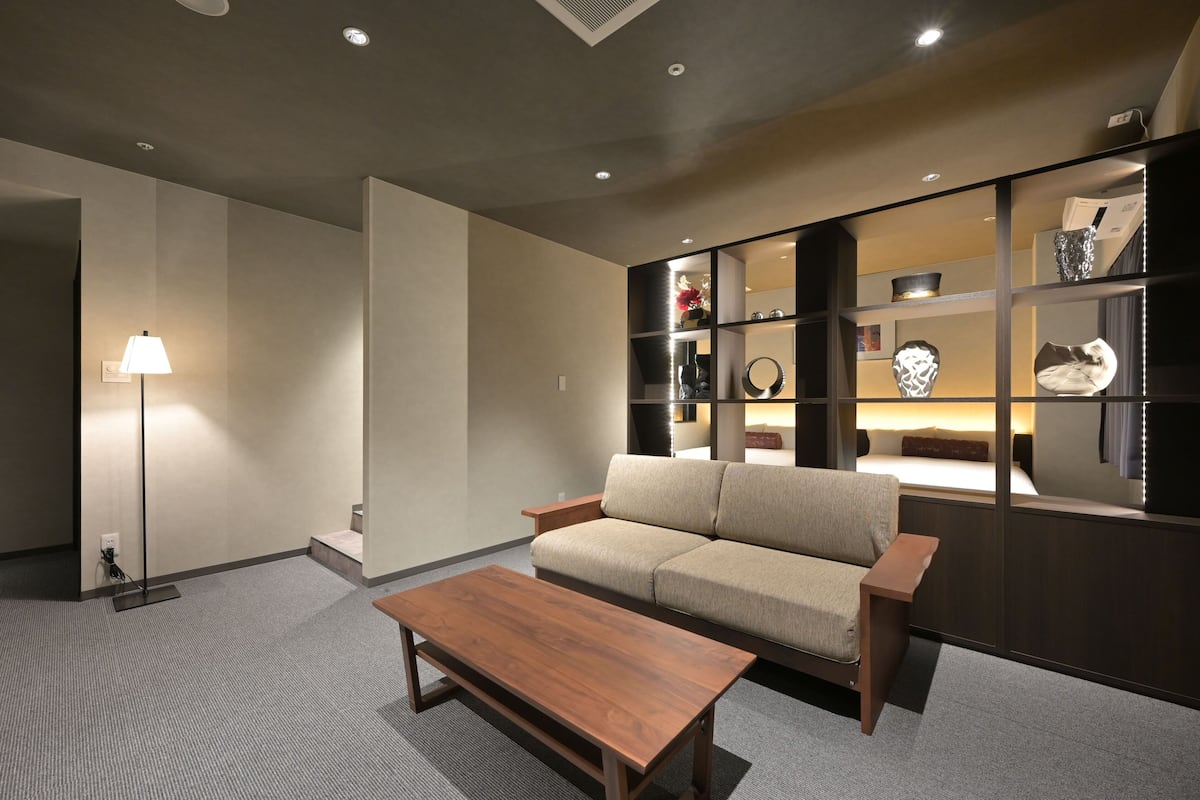A modern living room with a sofa, a wooden coffee table, and a floor lamp. There is a bookshelf with decorative items and a mirror.