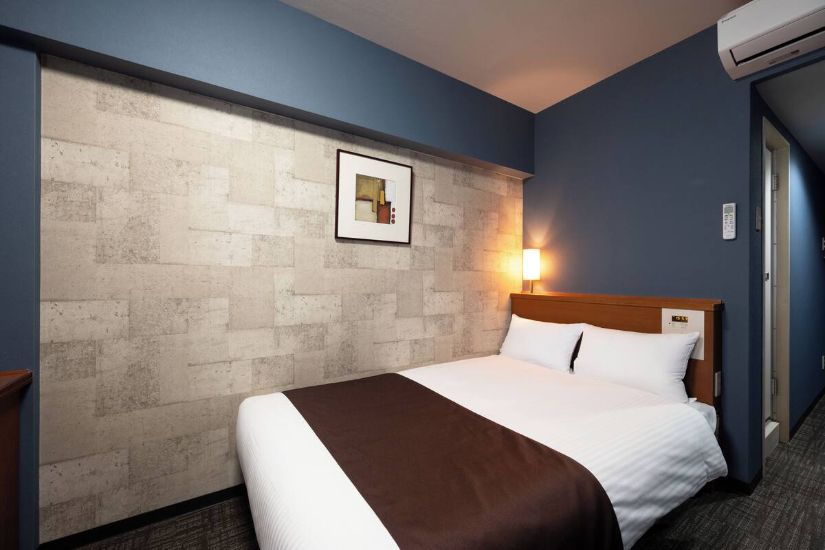 A hotel room with a large bed, a brick-patterned accent wall, a framed picture, a bedside lamp, and an air conditioning unit.