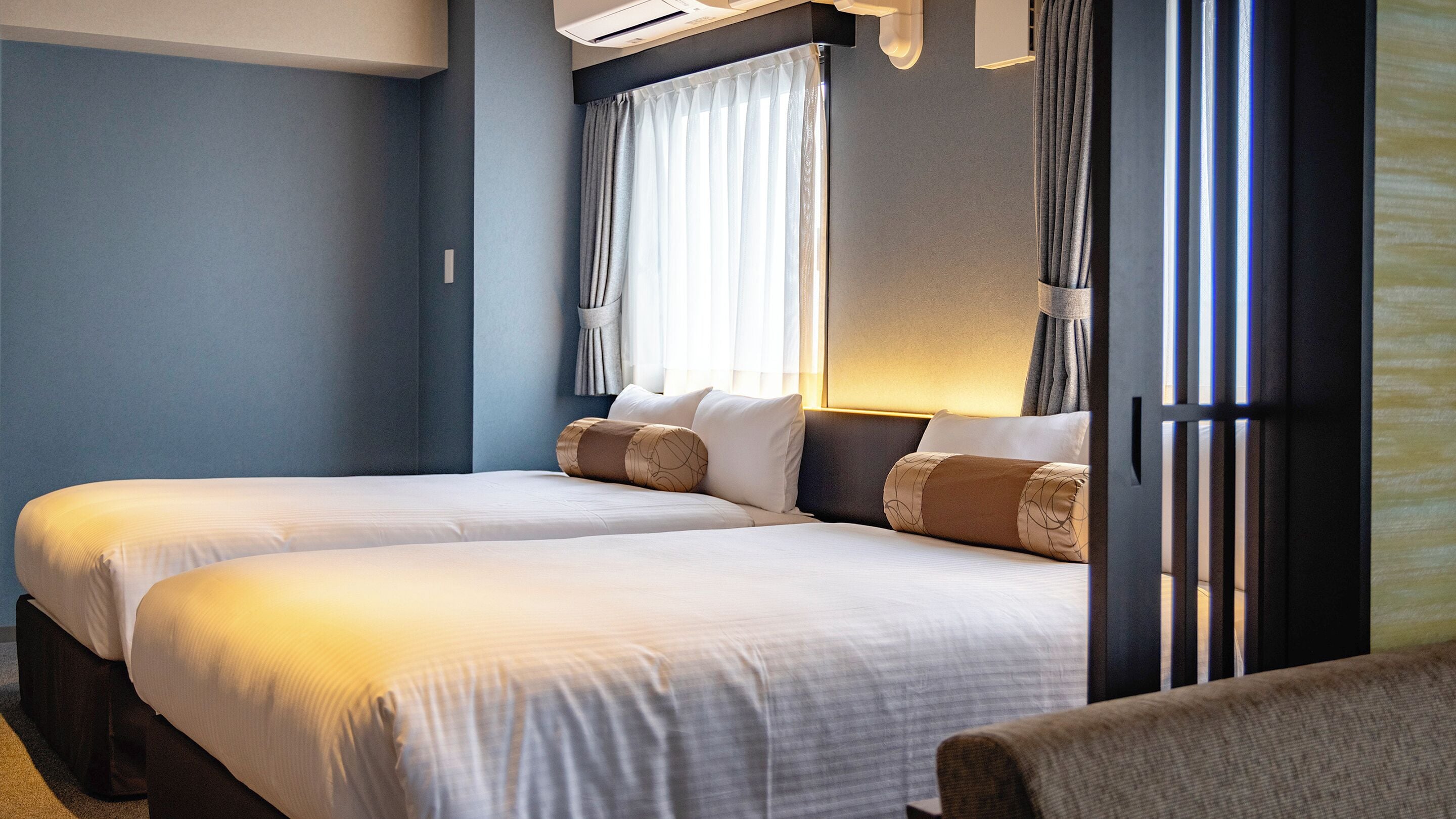 A hotel room with two beds, a large window with curtains, a wall-mounted air conditioner, and a bedside table.