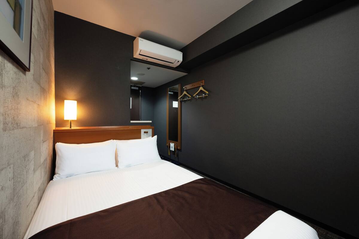 A hotel room with a bed, a wall-mounted air conditioner, a bedside lamp, and a mirror with a wooden hanger.