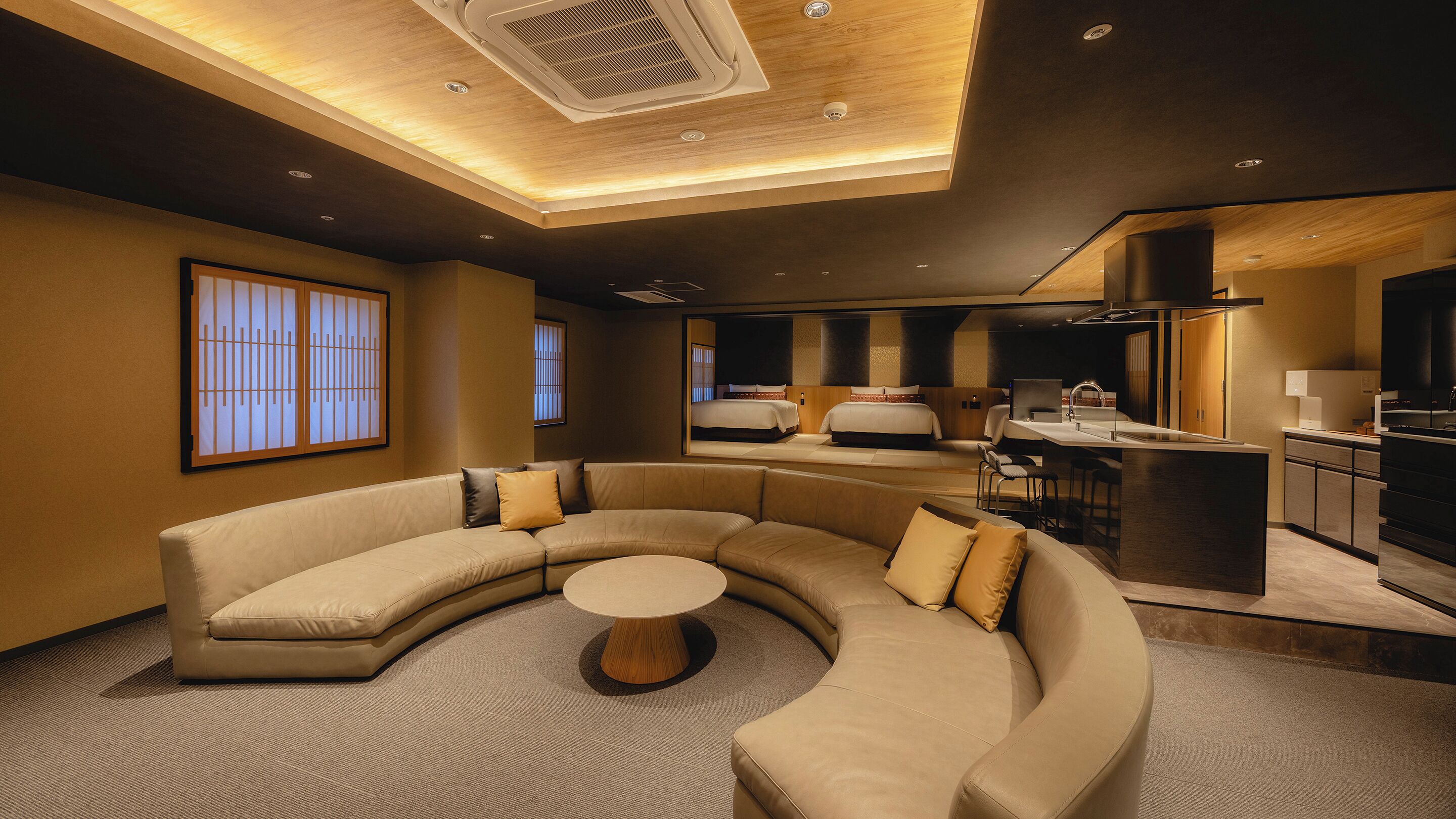 A modern hotel room with a curved sofa, a small round table, a kitchenette, and a large bed with a headboard.