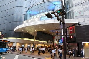 Una concurrida calle urbana con un edificio grande y curvo que dice "SHIMOTORI".