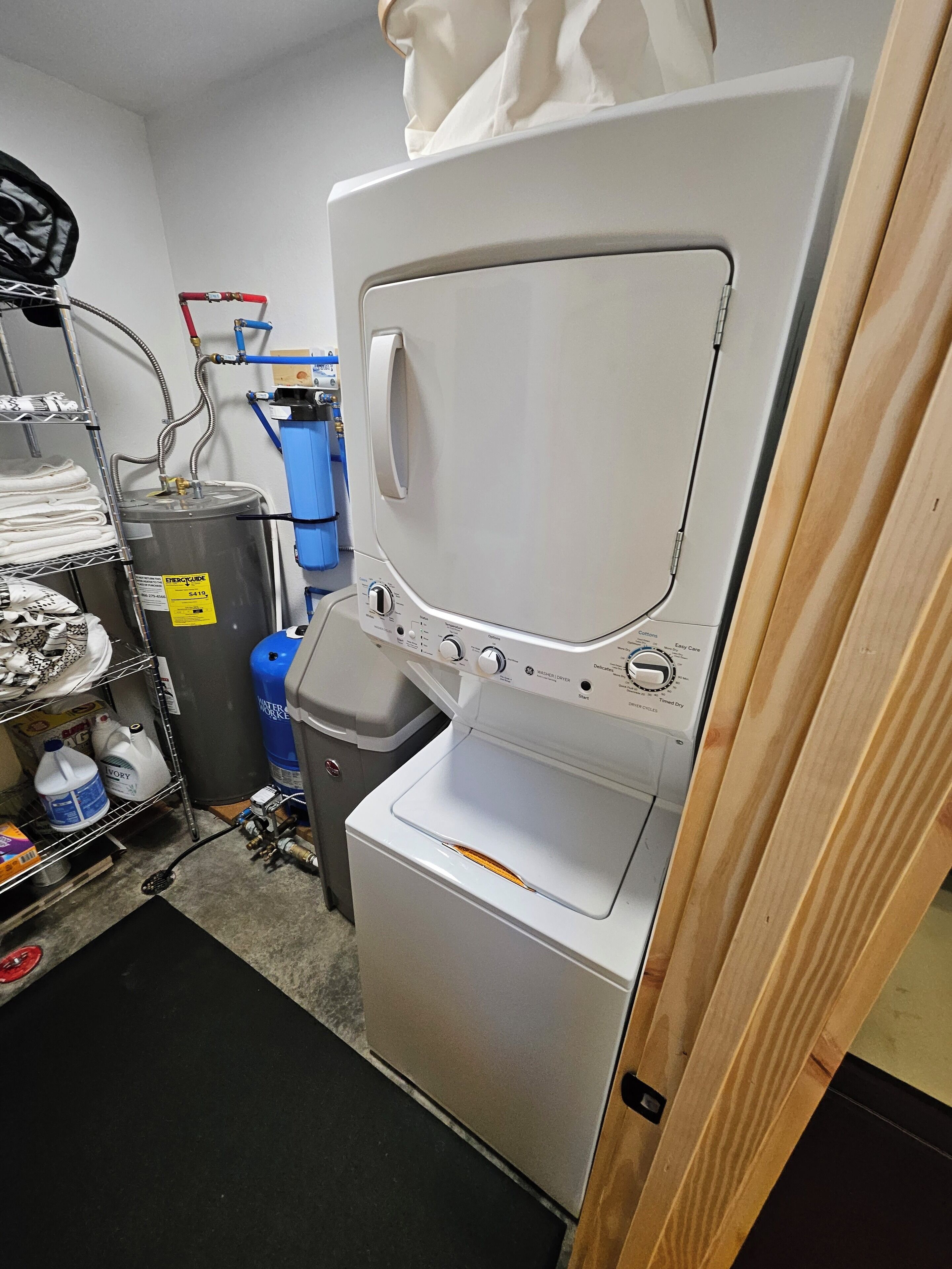 A laundry room with a stacked washer and dryer, a water heater, and various cleaning supplies.
