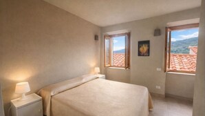 A bedroom with a bed, two bedside tables with lamps, two windows with a view of a tiled roof and greenery, and a framed picture on the wall. - Borgo Agapito in the heart of Marciana (Marciana)