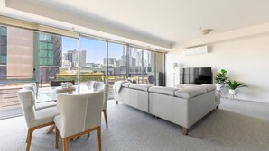 A modern living room with a sofa, dining table, and chairs, large windows offering a city view, a flat-screen TV, and a potted plant. - Vista on Queensbridge - 3BR Home in the Heart of It All (Southbank)