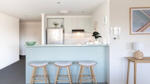 A modern kitchen with a light blue island, wooden stools, and white cabinetry. - Vista on Queensbridge - 3BR Home in the Heart of It All (Southbank)