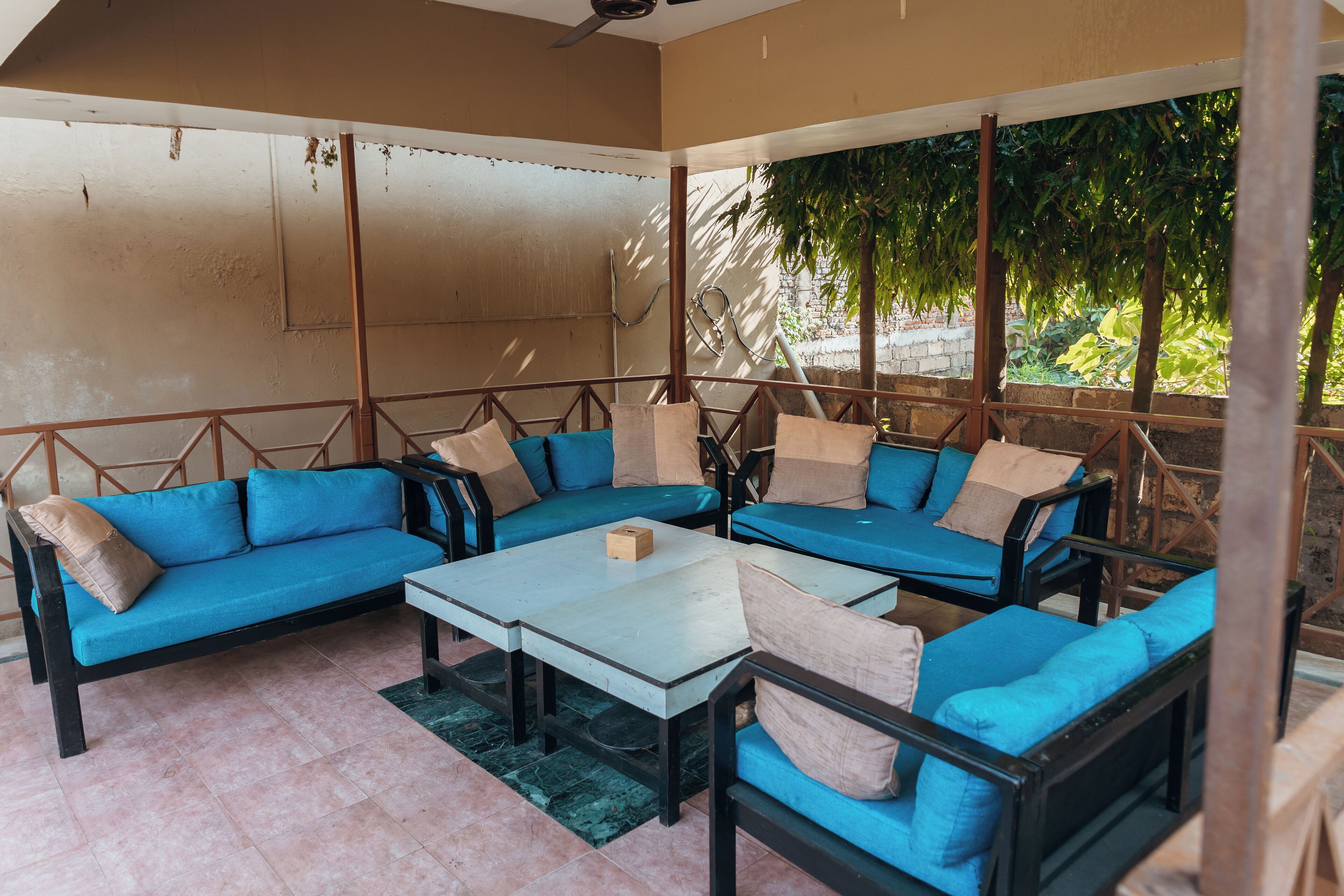 A covered outdoor seating area with blue sofas and a central table, surrounded by a wooden fence and a view of greenery.