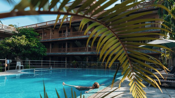 Ein Swimmingpool mit einer Person im Wasser, umgeben von Palmen und einem Gebäude mit Balkonen.
