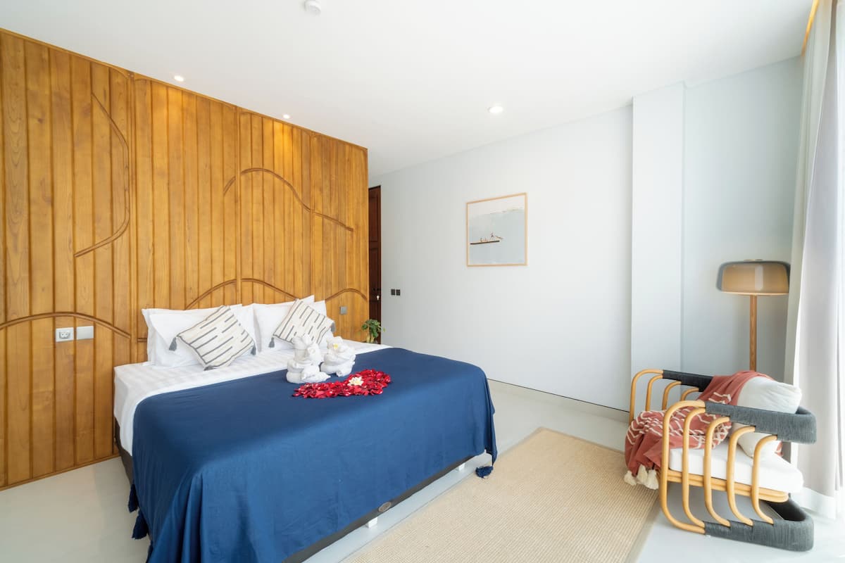 A modern bedroom with a wooden headboard, a blue bedspread, white bedding, and a small bedside table with a lamp.