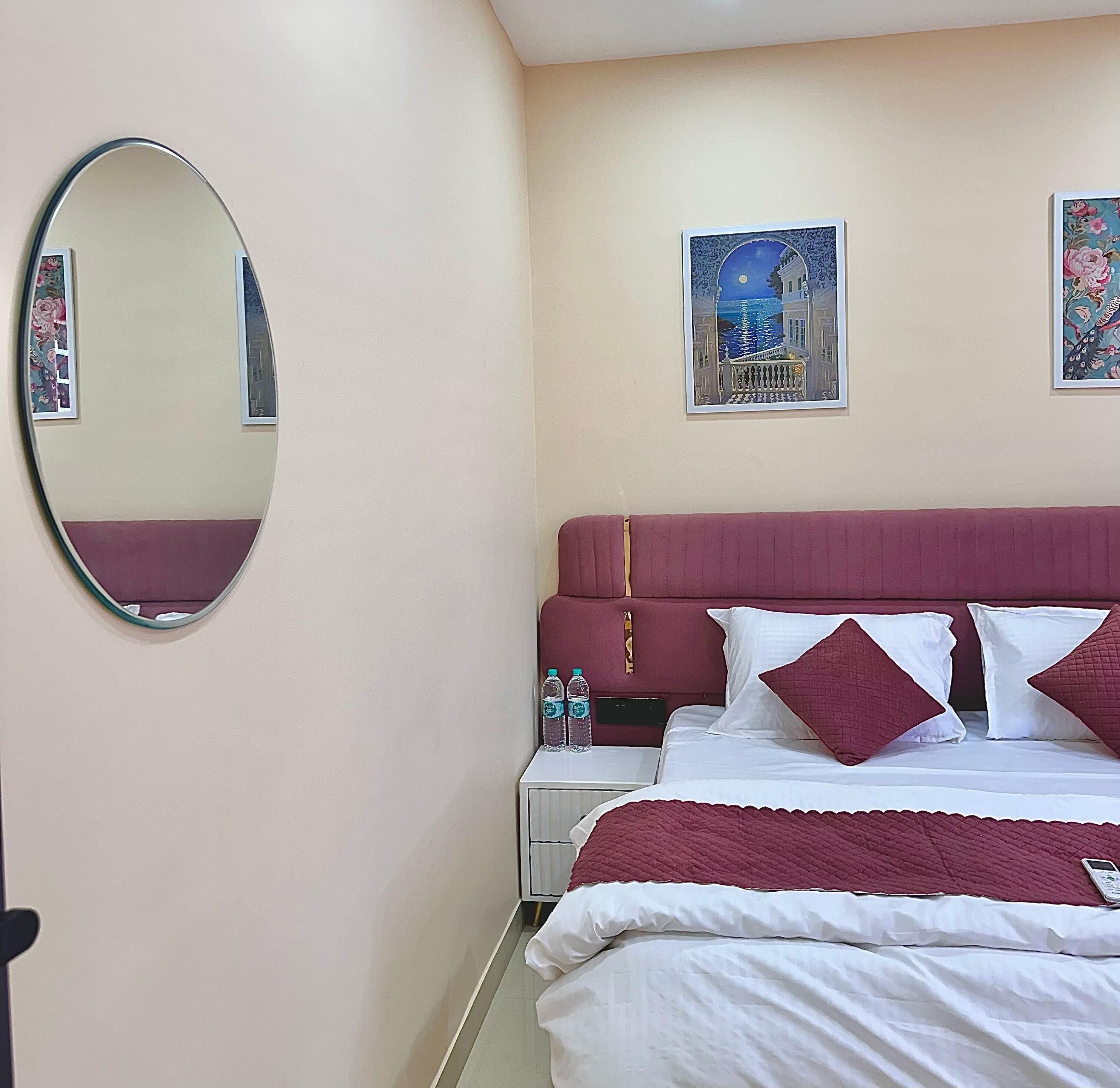 A hotel room with a large oval mirror, a bed with red bedding, two pillows, a nightstand with water bottles, and a framed picture on the wall.