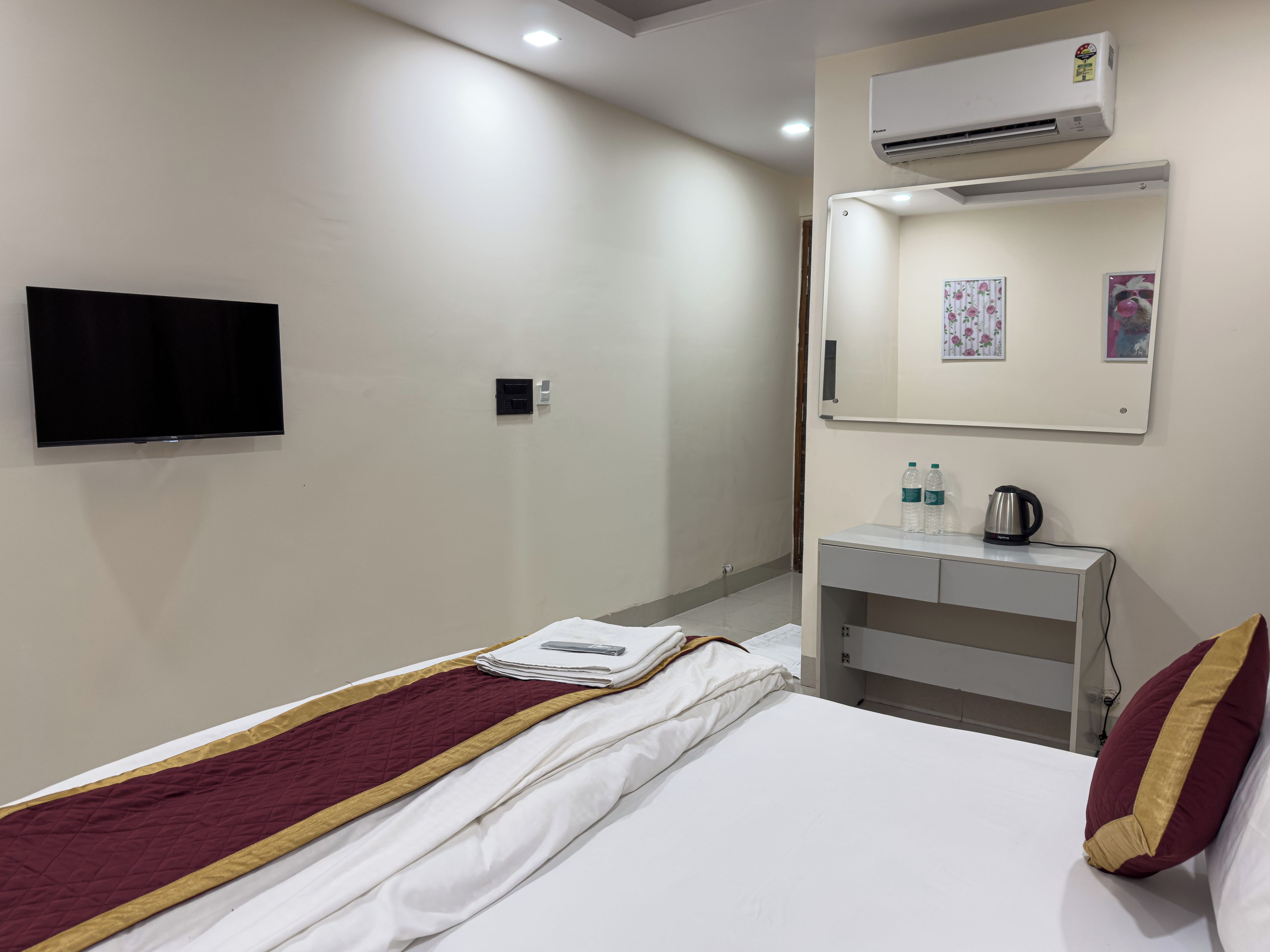A hotel room with a bed, a TV, an air conditioning unit, a desk with a kettle, and a mirror.