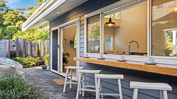 Un espace bar extérieur moderne, doté d’un comptoir en bois, de tabourets blancs et d’une grande fenêtre donnant sur un jardin.