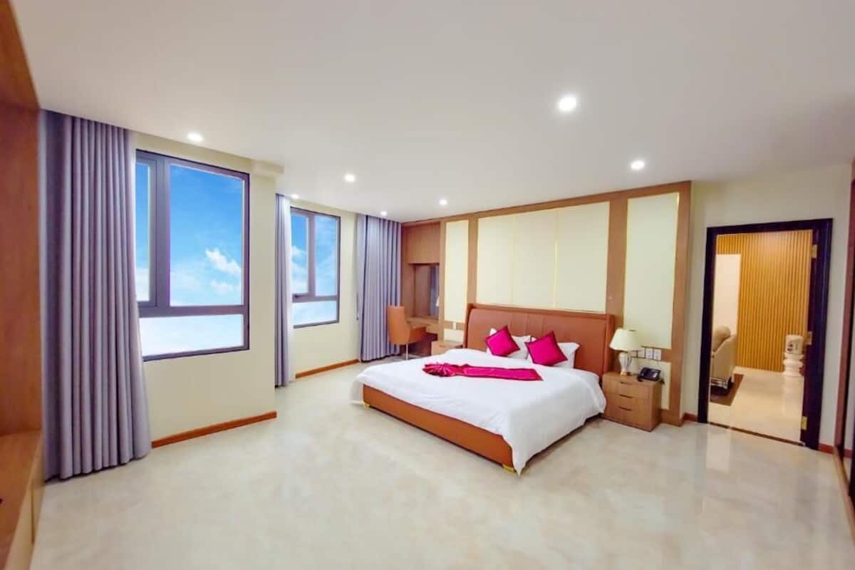 A spacious hotel room with a large bed, a desk, and a view of the sky through the windows.