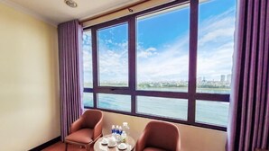 A room with a large window, two chairs, a small table with teapots and bottles, and a view of a city and water. - T.26 Hotel (Da Nang)