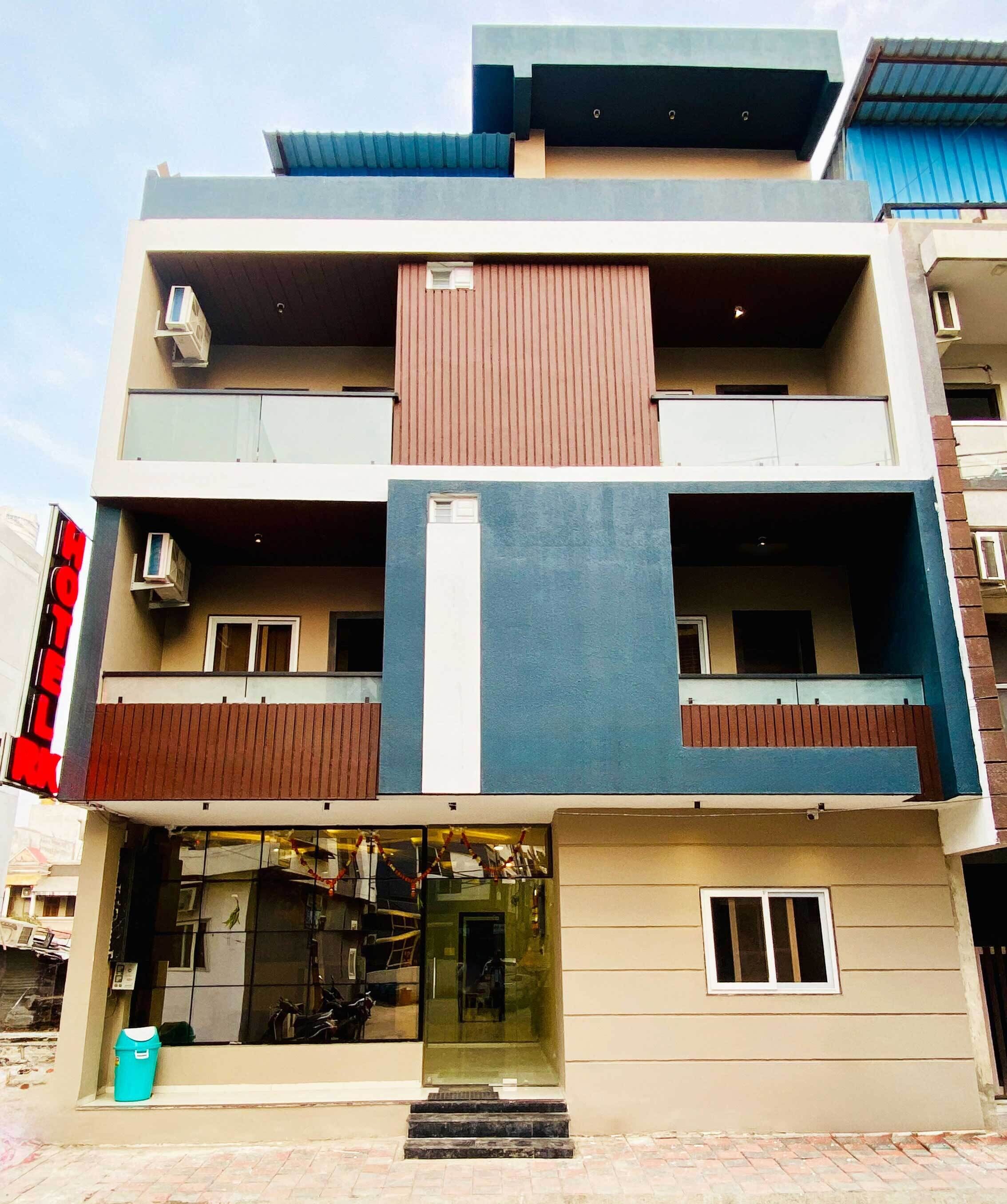 Un hotel de varios pisos con diseño moderno, que presenta una entrada central con puertas de vidrio, balcones en cada planta y unidades de aire acondicionado en el exterior.