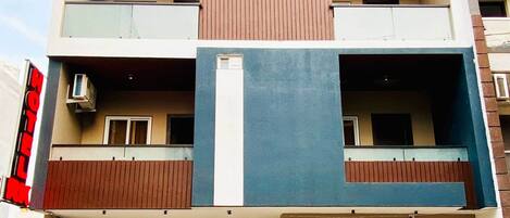Un hotel de varios pisos con diseño moderno, que presenta una entrada central con puertas de vidrio, balcones en cada planta y unidades de aire acondicionado en el exterior.