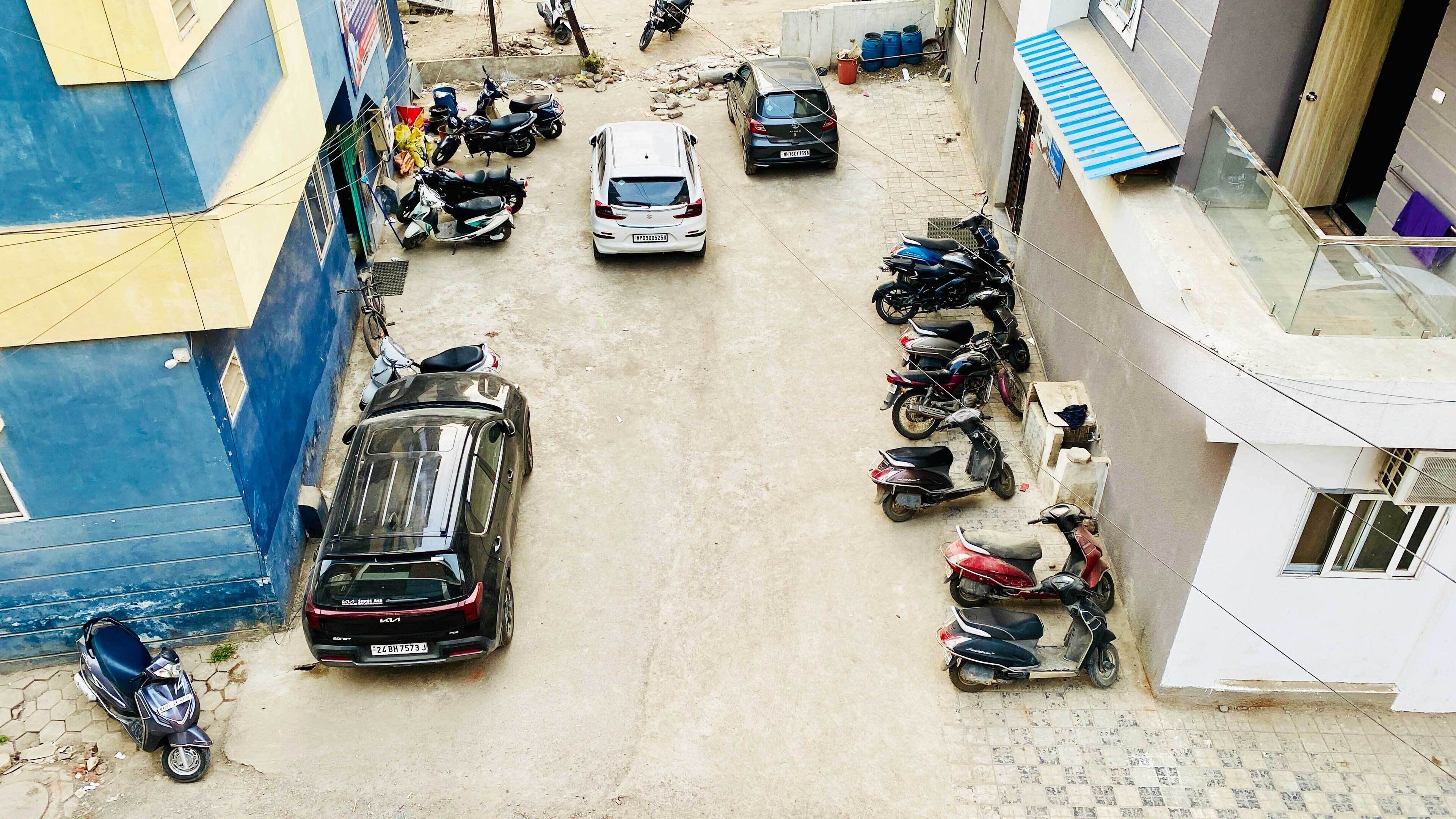 A narrow street with parked cars and scooters, buildings on either side, and a blue wall on the left.