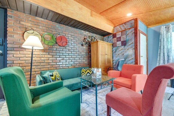 A cozy living room with a brick wall, a wooden ceiling, a green sofa, a red armchair, and a glass coffee table.