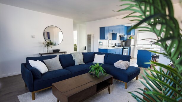 Un salon moderne avec un canapé d’angle bleu foncé, une table basse en bois et un tapis blanc. On aperçoit, au fond, une cuisine avec des meubles bleus.