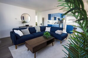Uma sala de estar moderna com um sofá seccional azul escuro, uma mesa de centro de madeira e um tapete branco. Uma cozinha com armários azuis é visível ao fundo.