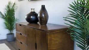 Un meuble en bois surmonté de deux vases, d’une plante en pot et d’un grand miroir accroché au mur.
