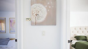 Un couloir avec une porte blanche, une œuvre d’art encadrée représentant un pissenlit, et une chambre adjacente avec un lit recouvert d’une literie verte.