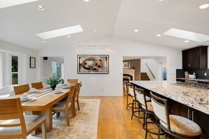 A spacious kitchen with a long island, wooden dining table, and chairs. There is a large painting on the wall and skylights on the ceiling.